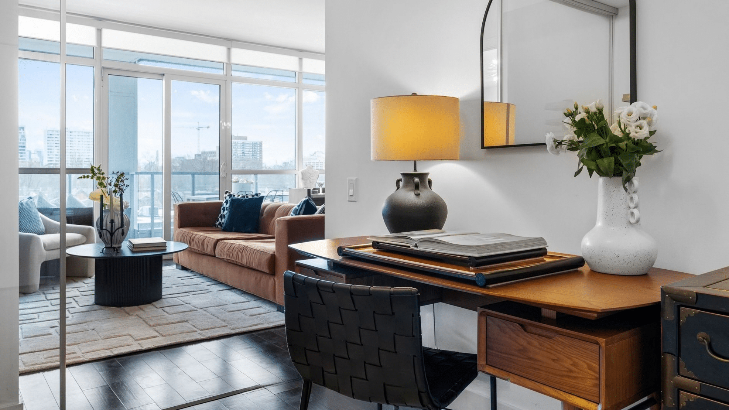 Modern home office with walnut desk, black woven chair, table lamp, artwork, books and adjacent living area